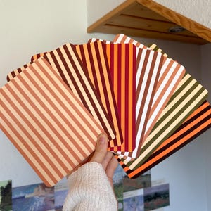 Könnte beinhalten: Ein Fächer aus rechteckigen Karten mit vertikalen Streifen in verschiedenen herbstlich inspirierten Farbpaletten, darunter Orange-, Braun-, Rot-, Creme- und Olivgrüntöne. Jede Karte zeigt ein durchgehendes Streifenmuster auf der Oberfläche.
