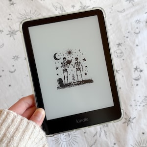 May include: A black Kindle e-reader with a clear protective case. The screen displays a black and white illustration of two skeletons, a crescent moon, stars, and a tombstone. The e-reader is held against a white patterned background.