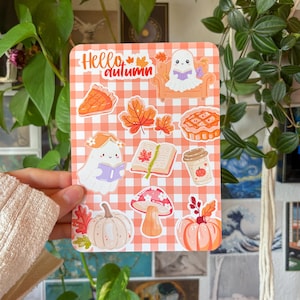 May include: A rectangular sticker sheet with an orange and white gingham background featuring autumn-themed illustrations including ghosts reading books, a pumpkin pie slice, a whole pie, maple leaves, a coffee cup, a mushroom, and pumpkins.