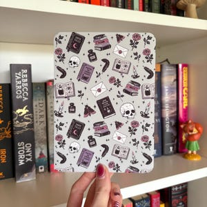 Könnte beinhalten: Ein graues Notizbuch mit einem sich wiederholenden Muster aus Totenköpfen, Büchern, Rosen, Federn und Tintenfässern. Das Notizbuch trägt die Aufschrift "DARK TALES" und "POETRY FOR CURSED HEARTS". Das Notizbuch wird vor einem Bücherregal gehalten.