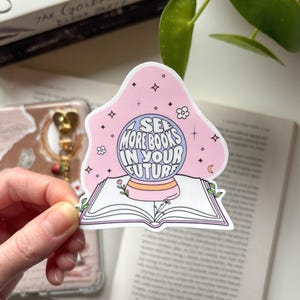 Könnte beinhalten: Ein rosa Aufkleber mit dem Text "SEE MORE BOOKS IN YOUR FUTURE" in einem Kristallkugel-Design. Der Aufkleber zeigt ein Buch, Blumen, Sterne und einen Mond. Der Aufkleber wird vor einem offenen Buch gehalten.