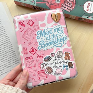 May include: A pink tablet case with a cow print design and decorative stickers. The stickers include books, hearts, and the text "Meet me at the Bookshop" and "the homebody club". A gold heart-shaped clasp is visible.