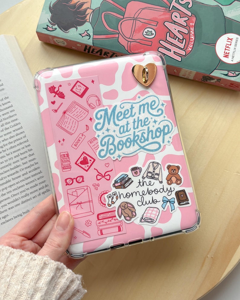 May include: A pink tablet case with a cow print design and decorative stickers. The stickers include books, hearts, and the text "Meet me at the Bookshop" and "the homebody club". A gold heart-shaped clasp is visible.