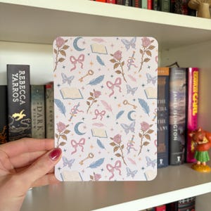 Könnte beinhalten: Eine rechteckige Karte mit einem sich wiederholenden Muster aus Rosen, Schmetterlingen, Schleifen, Schlüsseln, Federn, Monden und Büchern in Pastellfarben. Die Karte wird vor einem Bücherregal gehalten.