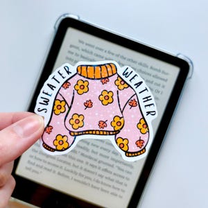 Könnte beinhalten: Ein rosa Aufkleber in Form eines Pullovers mit den Worten "SWEATER WEATHER" in einem Bogen. Der Pullover hat orangefarbene Blumen und Blätter sowie einen orangefarbenen Kragen und Bündchen. Der Aufkleber wird vor einem E-Reader gehalten.