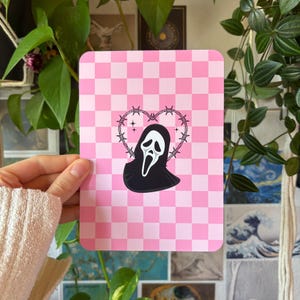 Puede incluir: Una impresión rectangular con un fondo de cuadros rosa claro y blanco. En el centro, una figura negra similar a un fantasma con una máscara blanca está enmarcada por un corazón hecho de alambre de espino con pequeñas estrellas de cuatro puntas.