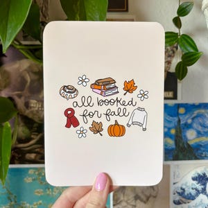 Könnte beinhalten: Eine weiße rechteckige Karte mit abgerundeten Ecken mit dem Text "all booked for fall" und Illustrationen eines Zimtschnecken, Bücher, Blätter, einem Kürbis und einem Pullover.