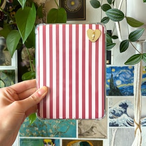 May include: A clear phone case with a red and white striped pattern. A gold heart-shaped charm is attached to the case. The case is rectangular and designed to fit a mobile phone.