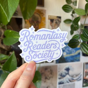 Könnte beinhalten: Ein hellvioletter Aufkleber mit dem Schriftzug "Romantasy Readers Society" in einer verspielten Schriftart. Der Aufkleber hat einen glitzernden, schillernden Effekt und ist mit kleinen Sternformen verziert. Der Aufkleber wird von einer Hand gehalten.