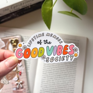 Könnte beinhalten: Ein farbenfroher Aufkleber in Wolkenform mit dem Text "LIFETIME MEMBER of the GOOD VIBES SOCIETY". Das Design verwendet orange, rosa, grüne und violette Buchstaben mit Blumen- und Smiley-Gesichtsdetails. Der Aufkleber wird in einer Hand gehalten.