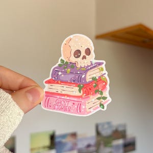 Op de afbeelding: Een gestanste sticker met een beige schedel die op een stapel van drie boeken rust. De boeken zijn paars, rood en roze, met groene klimop eromheen gewikkeld. Het onderste roze boek heeft de tekst GHOST STORIES op de voorkant gedrukt.