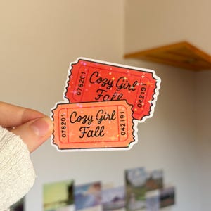 Könnte beinhalten: Zwei rechteckige Aufkleber in Form von Eintrittskarten mit gewelltem Rand. Der obere Aufkleber ist rot, der untere orange, beide mit einem holografischen Sternenmuster. Auf jedem Ticket steht der Text Cozy Girl Fall in schwarzer Schrift.