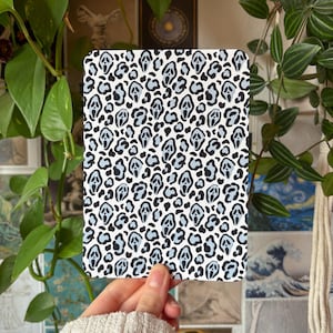 Op de afbeelding: Een rechthoekige kaart met een herhalend patroon van zwarte en lichtblauwe spookachtige maskers geïntegreerd in een luipaardprint. De achtergrond is wit en het patroon bestaat uit gestileerde schreeuwende gezichten omringd door donkere vlekken.