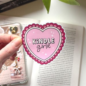 Könnte beinhalten: Ein rosa herzförmiger Aufkleber mit den Worten "KINDLE girlie" in schwarzer Schrift. Der Aufkleber hat einen gewellten Rand und kleine Sternakzente. Der Aufkleber wird vor einem offenen Buch gehalten.