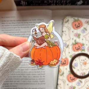 May include: A clear acrylic charm featuring a fairy with light brown hair and yellow wings sitting on a large orange pumpkin. The fairy is reading a brown book, accompanied by a small mouse, an acorn, and colourful autumn leaves at the base.