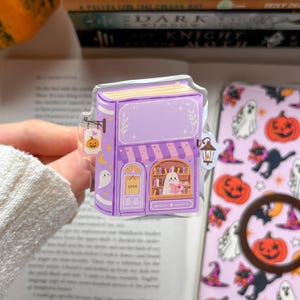 May include: A clear acrylic charm shaped like a purple bookstore with a striped awning, a ghost reading a book in the window, a hanging pumpkin sign, and a lantern. The charm features a small ghost, stars, and a crescent moon illustration.