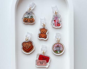 May include: Six clear acrylic charms with gold-tone jump rings and plastic clips. Designs include two pumpkins with a bow, a bear sitting on a pumpkin, a pink drink with a ghost, a heart cake labeled "I am an autumn", a gazebo, and two red tickets.