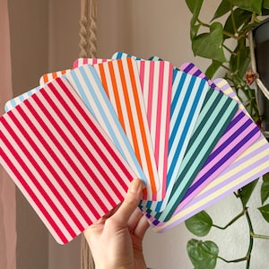 Könnte beinhalten: Eine Hand hält einen Fächer aus farbenfrohen, gestreiften Karten. Die Karten weisen vertikale Streifen in verschiedenen Farben auf, darunter Rot, Pink, Orange, Blau, Grün und Lila. Die Karten sind rechteckig mit abgerundeten Ecken.