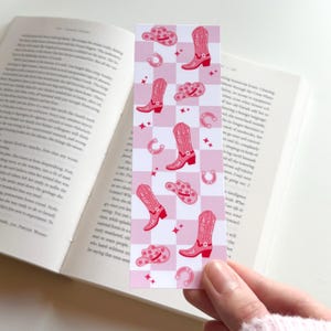May include: A pink and white chequered bookmark with illustrations of cowboy boots, hats, horseshoes, and stars. The bookmark is rectangular and held in front of an open book.