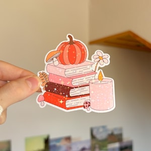 May include: A die-cut sticker featuring a stack of four books in shades of pink, brown, and red, topped with an orange pumpkin. A pink candle, a small daisy, and several small mushrooms are arranged around the books, all with a sparkly effect.