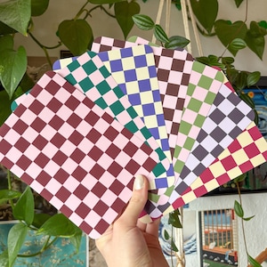 Può includere: Una mano tiene un ventaglio di carte colorate con motivi a scacchi. Le carte presentano una varietà di colori, tra cui rosa, bordeaux, verde, viola, marrone e giallo. Le carte sono rettangolari con angoli arrotondati.