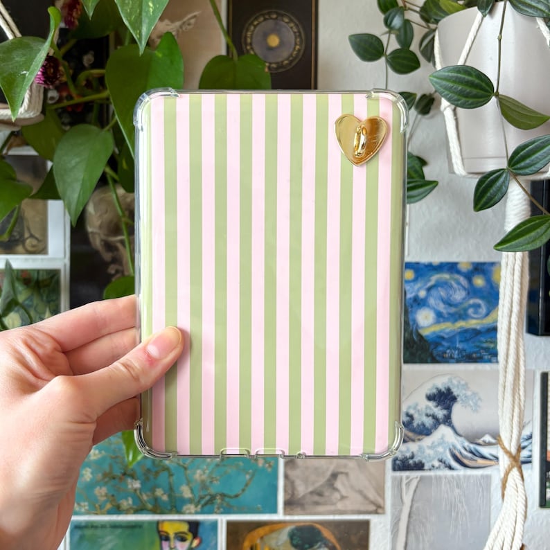 May include: A clear protective case for an e-reader with a vertical striped pattern in light pink and green. A gold heart-shaped charm is attached to the case. The case is held by a hand.