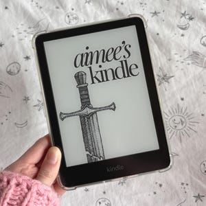 May include: A black Kindle e-reader with a clear protective case displays the text "aimee's kindle" above a detailed illustration of a sword. The word "kindle" is also visible at the bottom of the screen.