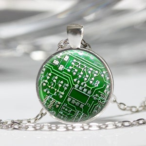 May include: A silver-toned necklace featuring a round pendant with a green circuit board design. The pendant showcases intricate white lines and components, resembling a miniature electronic circuit. The necklace chain is also silver.