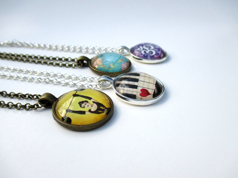 May include: Four round pendant necklaces with silver and bronze chains. The pendants feature diverse imagery including a yellow illustration of a person lifting weights, a piano keyboard with a red heart, a blue world map, and a purple Om symbol.