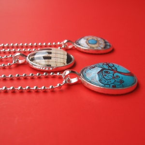 May include: Three silver-toned pendant necklaces with ball chains. One pendant features a black and white piano key design, another displays a blue tree illustration, and the third shows a detailed eye graphic under a clear glass dome.