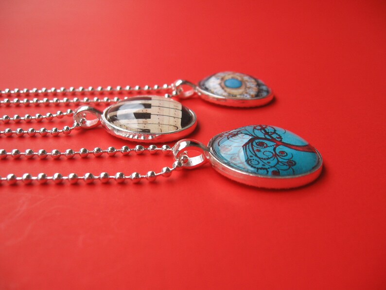 May include: Three silver-toned pendant necklaces with ball chains. One pendant features a black and white piano key design, another displays a blue tree illustration, and the third shows a detailed eye graphic under a clear glass dome.