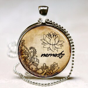 May include: A round silver pendant necklace featuring a beige background with a black lotus flower illustration, intricate floral scrollwork, and the word namaste written in a cursive script. The pendant hangs from a silver ball chain necklace.