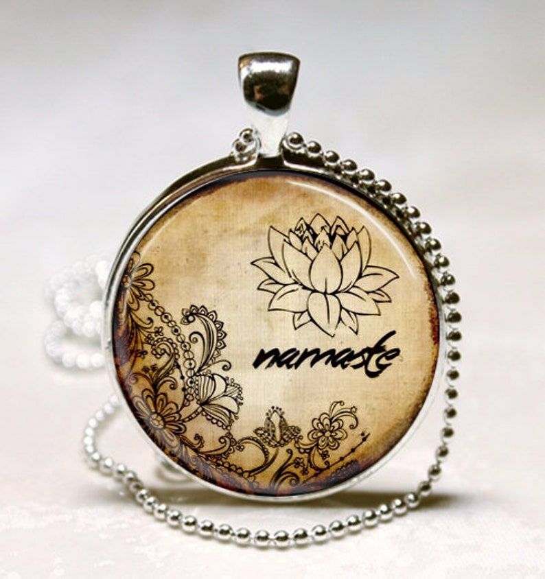 May include: A round silver pendant necklace featuring a beige background with a black lotus flower illustration, intricate floral scrollwork, and the word namaste written in a cursive script. The pendant hangs from a silver ball chain necklace.
