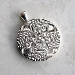 May include: A circular silver-toned pendant featuring a textured, dimpled surface on the front and a smooth, polished bail at the top. The metal has a reflective finish, and the circular face displays a consistent, fine-grained pattern throughout.