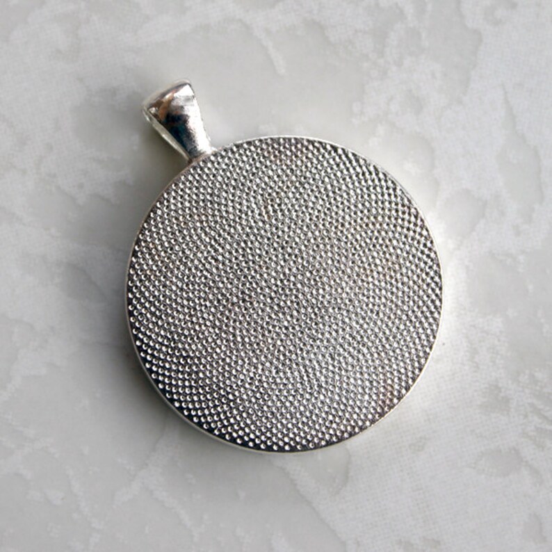 May include: A circular silver-toned pendant featuring a textured, dimpled surface on the front and a smooth, polished bail at the top. The metal has a reflective finish, and the circular face displays a consistent, fine-grained pattern throughout.