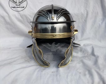 May include: A polished steel Roman helmet with brass accents. The helmet features a rounded top, a brow guard, and cheek pieces. The design includes decorative elements and is set against a white background. The logo "Eagle steel craft" is visible.
