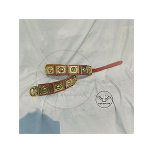 May include: A brown leather belt with brass-colored square plates and a matching buckle. The plates have a circular design in the center. The belt is lying on a white surface.