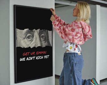May include: Black framed art print with a black background, featuring a torn image of a hundred-dollar bill with eyes visible. The text "GET UP, EMMA! WE AIN'T RICH YET" is written in red and white. A person is hanging the art.