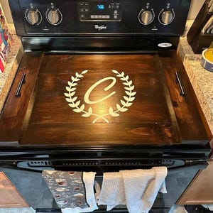 Könnte beinhalten: Eine dunkel gebeizte Holzherdabdeckung mit einem weißen Lorbeerkranz-Design und dem Buchstaben "C" in der Mitte. Die Abdeckung hat zwei schwarze Griffe und ist auf einem schwarzen Whirlpool-Herd platziert.