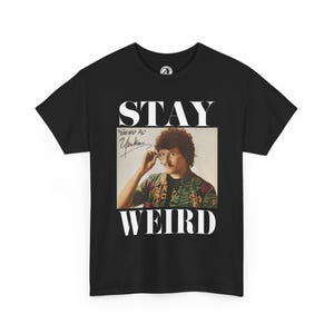 Puede incluir: Camiseta negra con las palabras "STAY WEIRD" en letras blancas. La camiseta presenta una foto de "Weird Al" Yankovic, con su firma. La imagen es de estilo vintage.