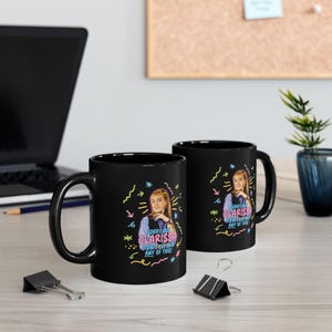 May include: Two black ceramic mugs with a colorful graphic of a woman and the text "Sorry but Clarissa didn't explain any of this!". The mugs have a glossy finish and a curved handle.