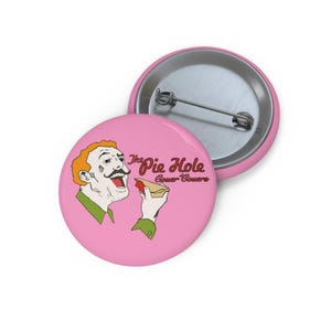 May include: Pink button with a cartoon illustration of a man eating pie, with the text "The Pie Hole Gower Couere". The man has orange hair and a mustache. The button is round and has a metal pin back.