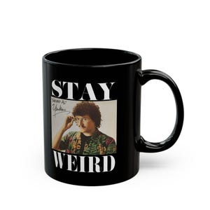 May include: Black ceramic mug featuring a portrait of "Weird Al" Yankovic with the words "STAY WEIRD" in white. The mug has a curved handle and a glossy finish, perfect for coffee or tea.