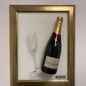 May include: A framed display featuring a bottle of Moët & Chandon Imperial Brut champagne and a champagne flute. The bottle is green with a gold foil label and a gold foil top. The frame is gold.