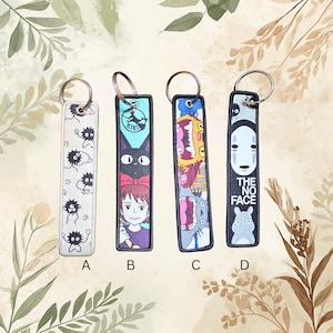 May include: Four rectangular fabric keychains with metal rings. Keychain A is white with black soot sprites. Keychain B features a girl, a black cat, and the text KIKI. Keychain C shows colorful monster faces. Keychain D is black with a white mask and text THE NO FACE.