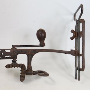 May include: A vintage cast iron hand-cranked kitchen tool featuring a dark brown rusted finish, a wooden handle knob, a gear mechanism, and a vertical mounting bracket with a wing nut for securing the device to a flat surface edge.