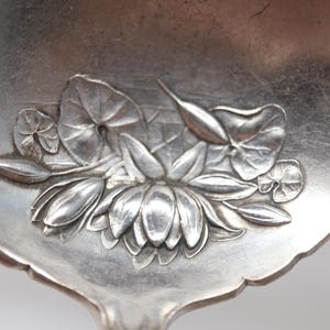 May include: A silver-toned metal spoon bowl featuring an embossed floral design. The relief pattern depicts a central water lily blossom surrounded by rounded lily pads and slender buds, all rendered in a polished metallic finish with fine details.