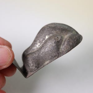 May include: A close-up of a small, antique, silver-colored metal object. The object has a curved, scoop-like shape with a flat base. The metal has a worn, textured appearance, suggesting age and use. The object is held in a hand.