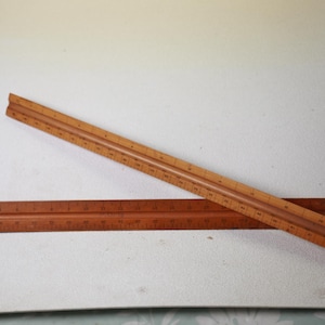 May include: Two wooden architectural rulers, one angled across the other. The rulers are light brown with dark brown markings. The top ruler is angled across the bottom ruler. The rulers are used for measuring and drafting.