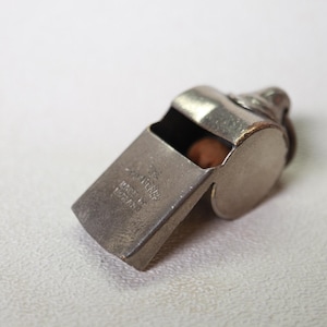 May include: A vintage silver-toned whistle with a rounded body and a rectangular mouthpiece. The whistle has visible wear and tear, suggesting age. The text "Made in England" is stamped on the side. A small, round, brown object is visible inside the whistle.